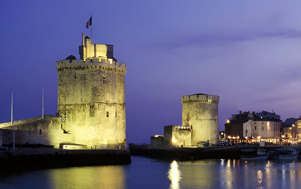 Towers of La Rochelle illuminated at dusk, reflecting on the water.
