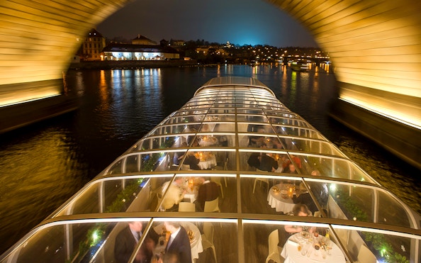 Open-top glass boat on Prague dinner cruise with live music, passing under a lit bridge at night.