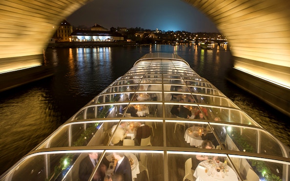 Open-top glass boat on Prague dinner cruise with live music, passing under a lit bridge at night.