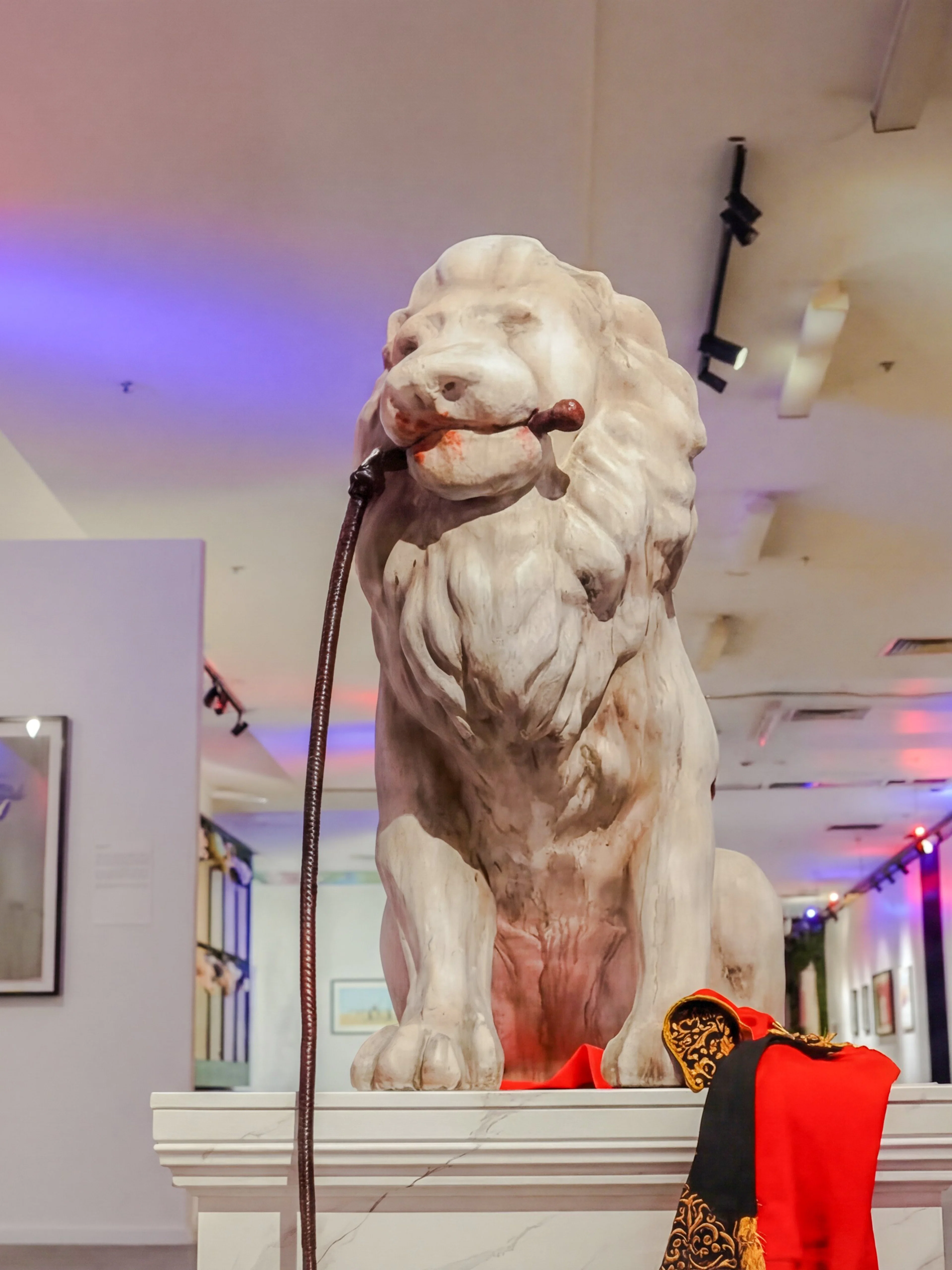 Lion statue with a leash in a Banksy art exhibit, surrounded by framed artworks.