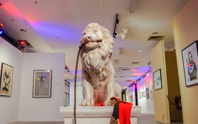 Lion statue with a leash in a Banksy art exhibit, surrounded by framed artworks.