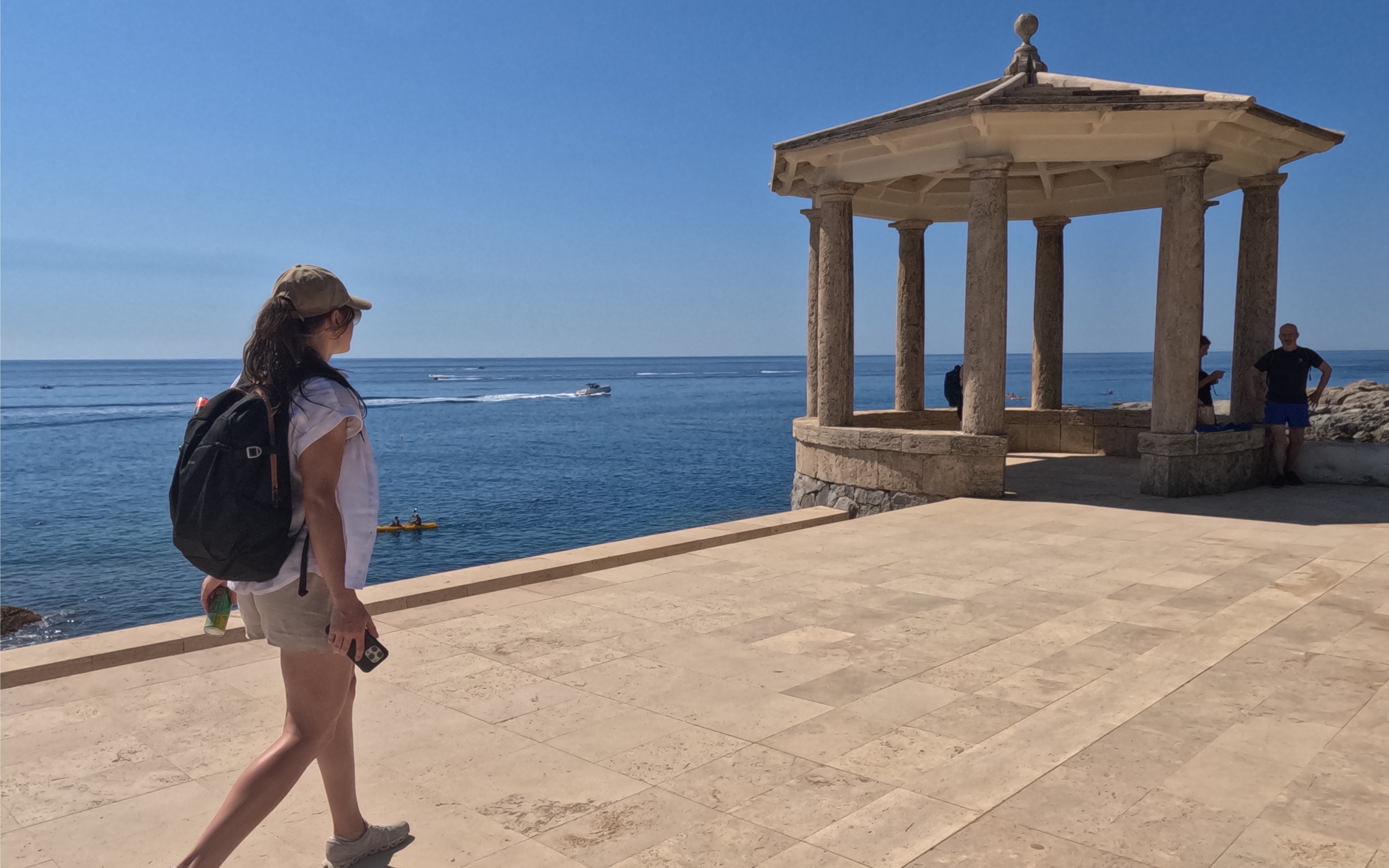 Woman walking on Camí de Ronda coastal path near a stone gazebo in S'Agaró, Spain.