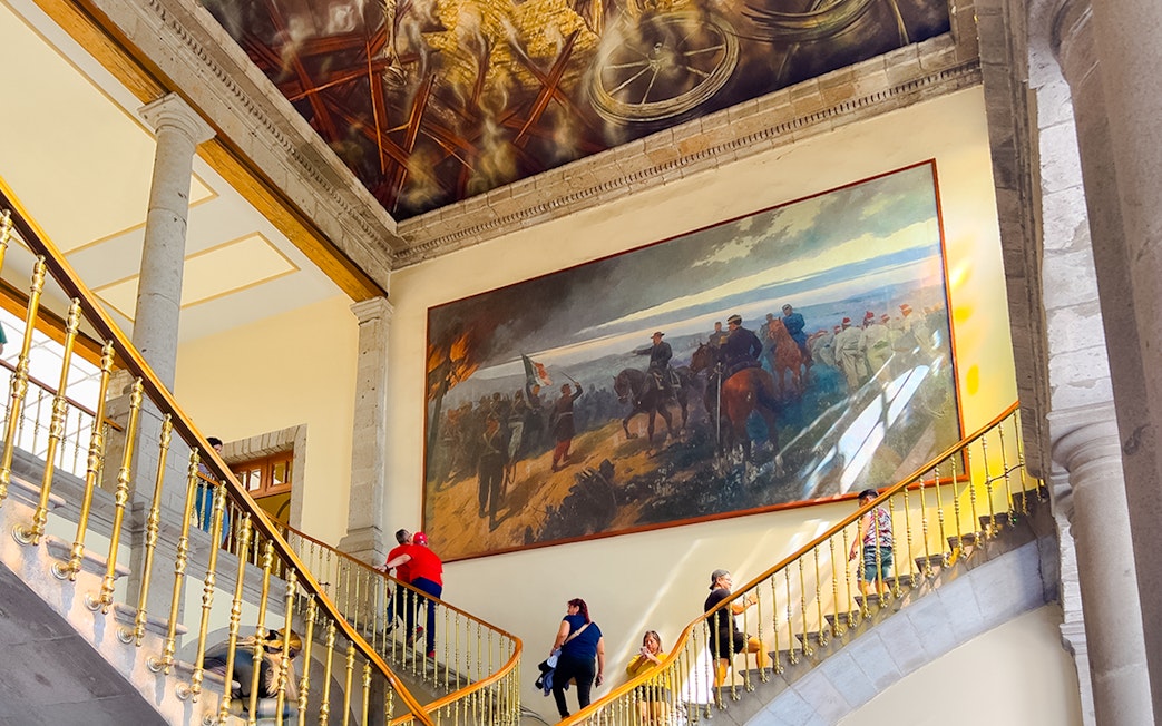 Visitors ascending staircase at Chapultepec Castle, Mexico, with historical mural and ornate ceiling.