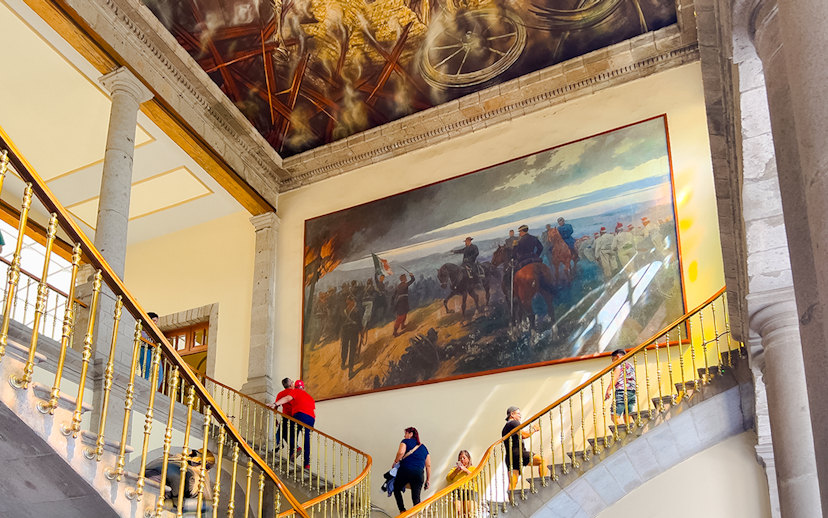 Visitors ascending staircase at Chapultepec Castle, Mexico, with historical mural and ornate ceiling.