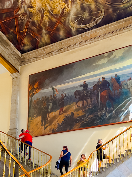 Visitors ascending staircase at Chapultepec Castle, Mexico, with historical mural and ornate ceiling.