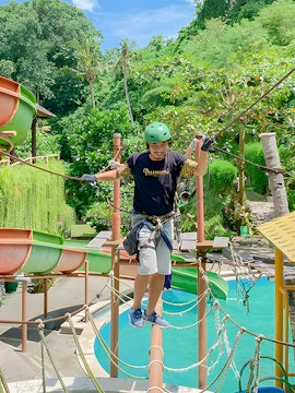 Person crossing a rope bridge at Keramas Park adventure course, surrounded by lush greenery.