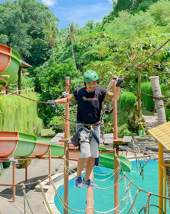 Person crossing a rope bridge at Keramas Park adventure course, surrounded by lush greenery.