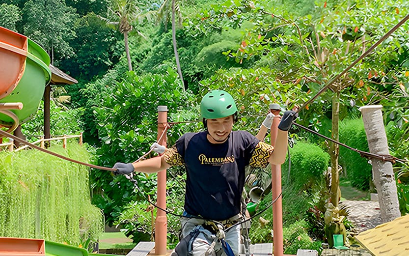 Person crossing a rope bridge at Keramas Park adventure course, surrounded by lush greenery.