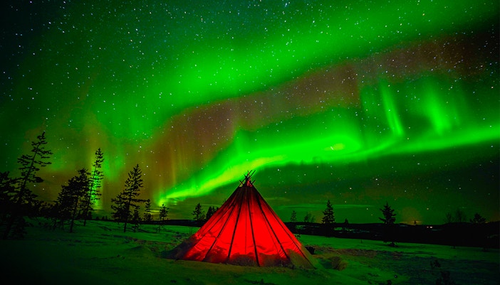 Northern Lights over a glowing tent in snowy Rovaniemi, Finland.