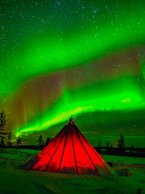 Northern Lights over a glowing tent in snowy Rovaniemi, Finland.