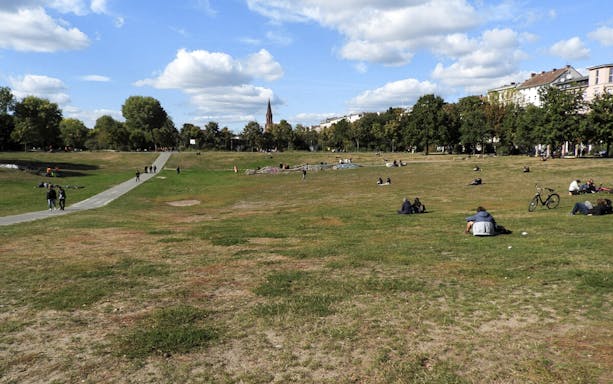 People relaxing in a grassy park in Kreuzberg, Berlin, with a church spire in the background.