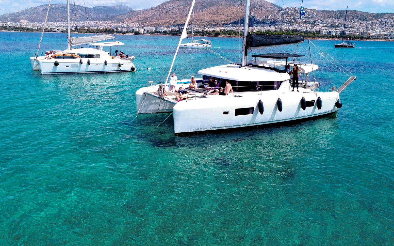 Lagoon 42 catamaran with people sailing in clear blue waters near a coastal city.