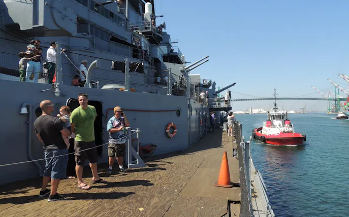 Visitors exploring a historic battleship docked at a harbor with a bridge in the background.