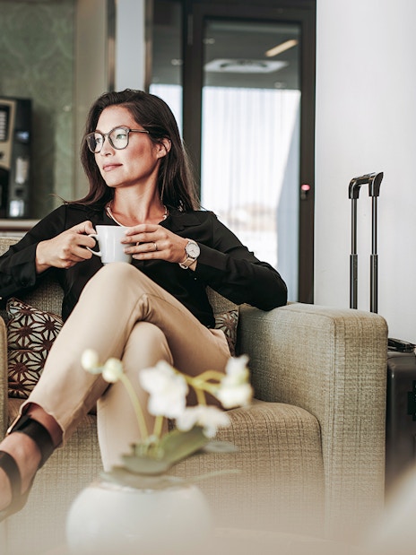 Person relaxing with coffee in an airport lounge, suitcase nearby.
