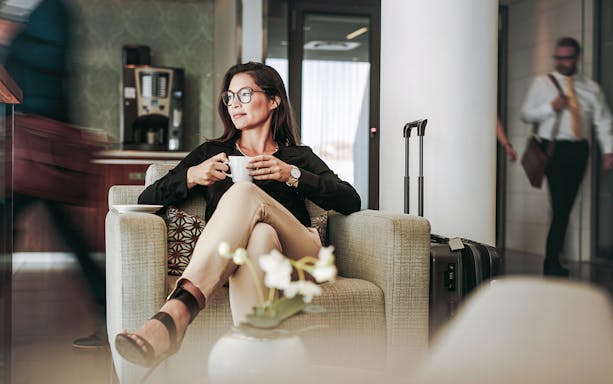 Person relaxing with coffee in an airport lounge, suitcase nearby.