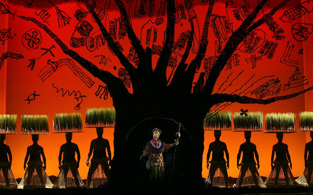 Stage performance of The Lion King with actors in traditional costumes and vibrant backdrop.