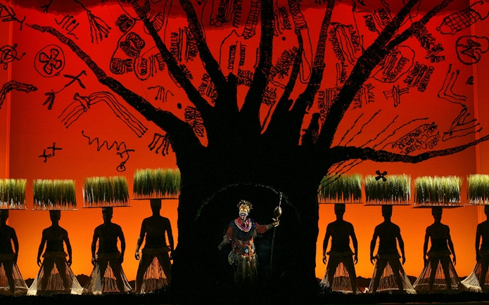 Stage performance of The Lion King with actors in traditional costumes and vibrant backdrop.