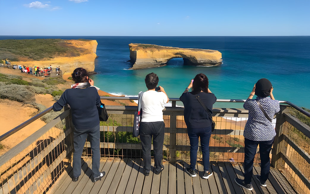 Tourists at a viewpoint capturing the scenic view of the Great Ocean Road's coastal arch formation.