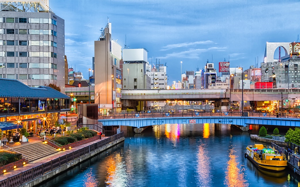 Tombori River Cruise boat on Dotonbori Canal in Osaka, Japan, with cityscape and illuminated buildings.