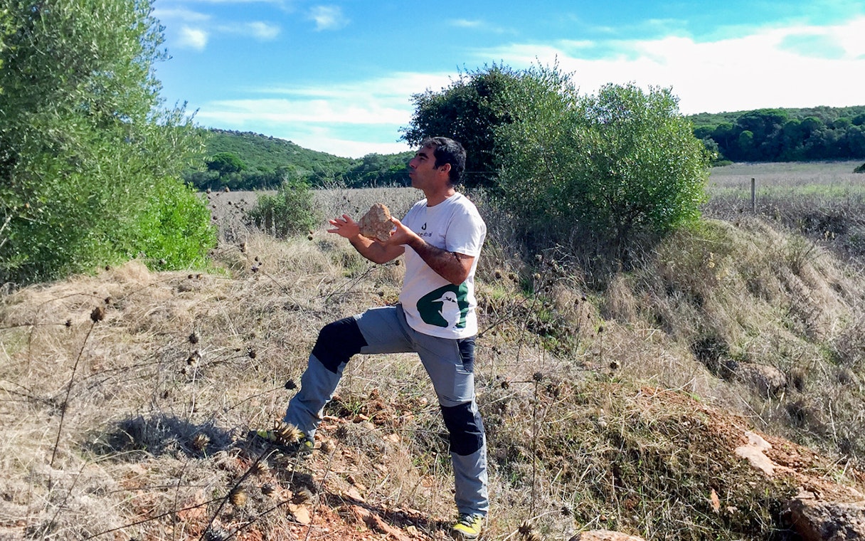 Man hiking in Arrábida Natural Park, Portugal, holding a rock amidst greenery and open fields.
