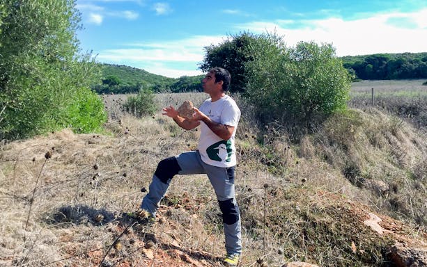 Man hiking in Arrábida Natural Park, Portugal, holding a rock amidst greenery and open fields.