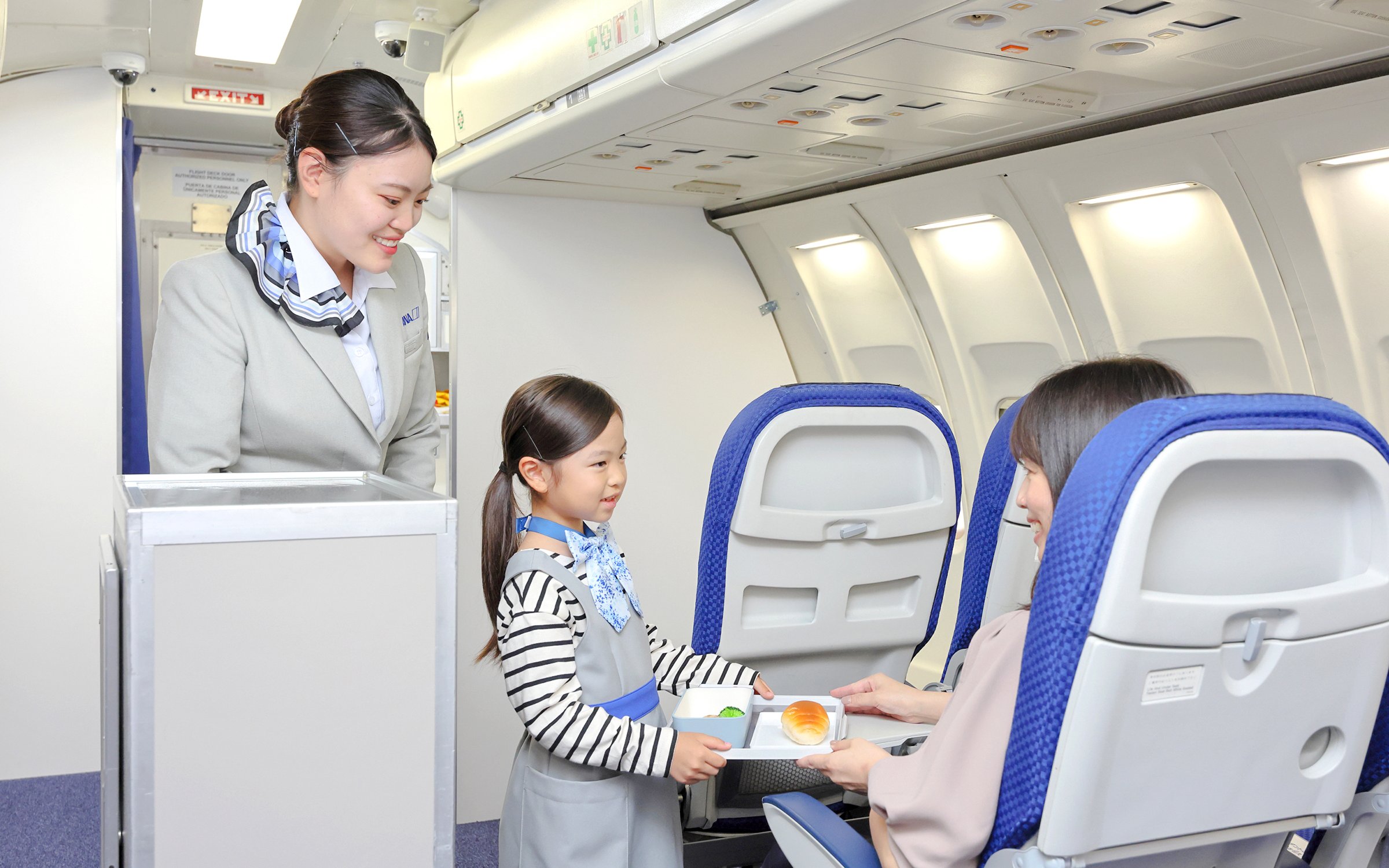 Child serving food on airplane at KidZania Fukuoka with air hostess assisting.