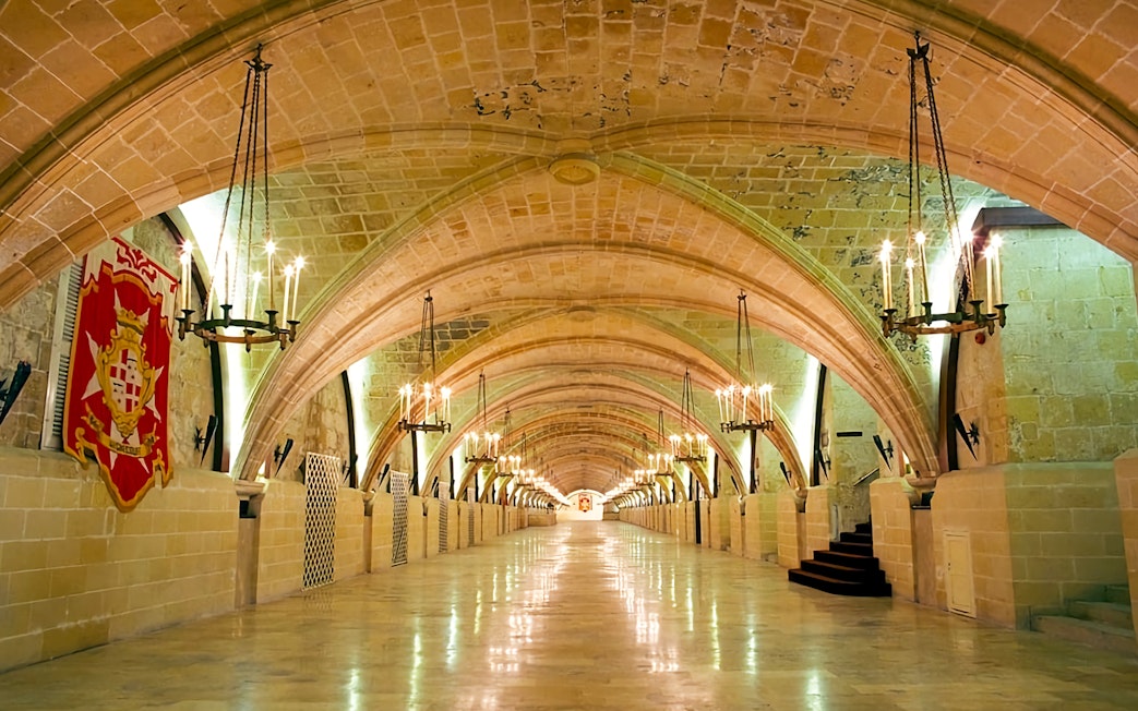 Grand arched hallway in the Holy Infirmary, Malta, with chandeliers and a Maltese cross banner.