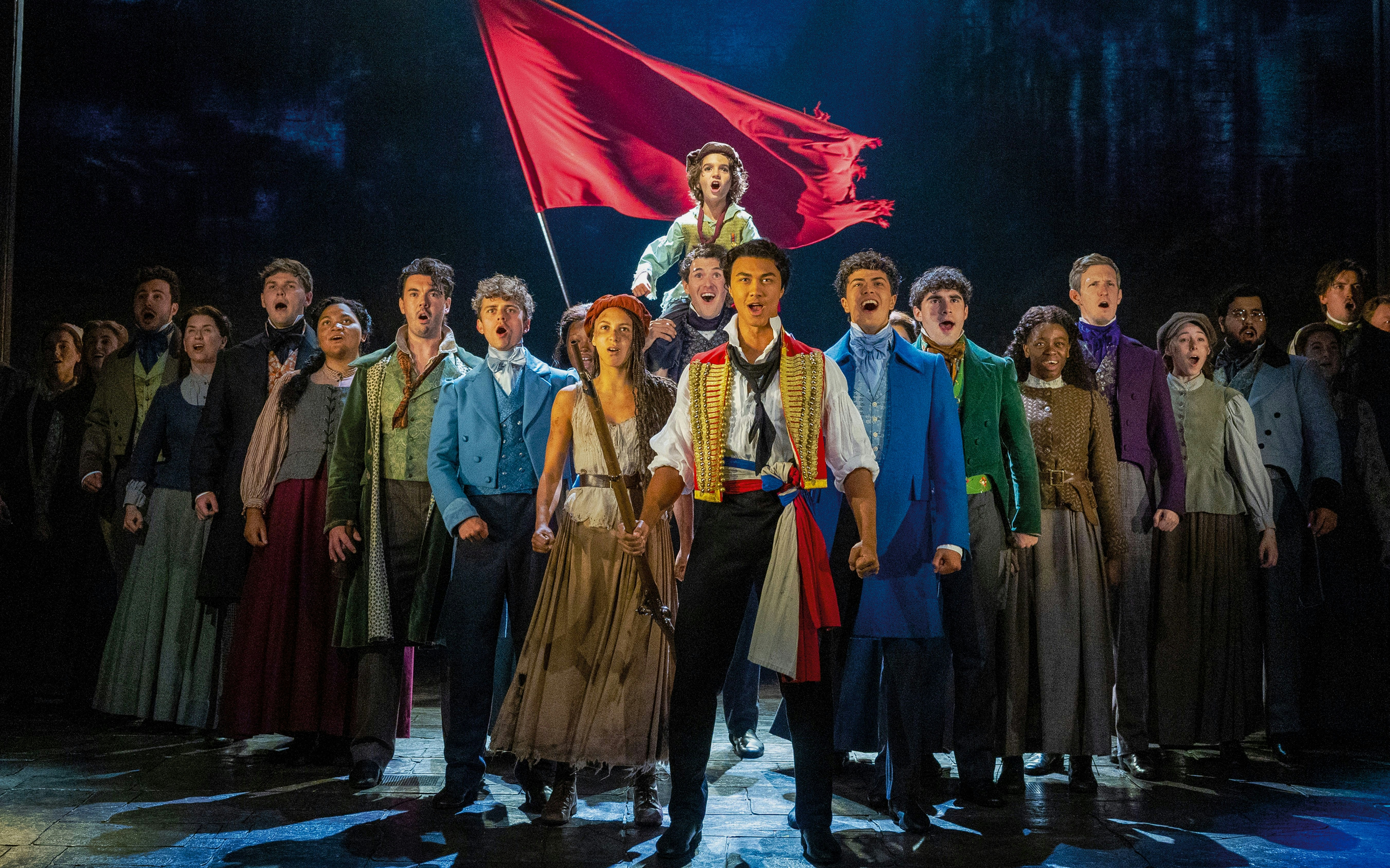 Les Misérables cast performing on stage with a red flag in the background.