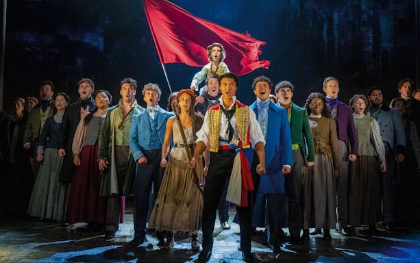 Les Misérables cast performing on stage with a red flag in the background.