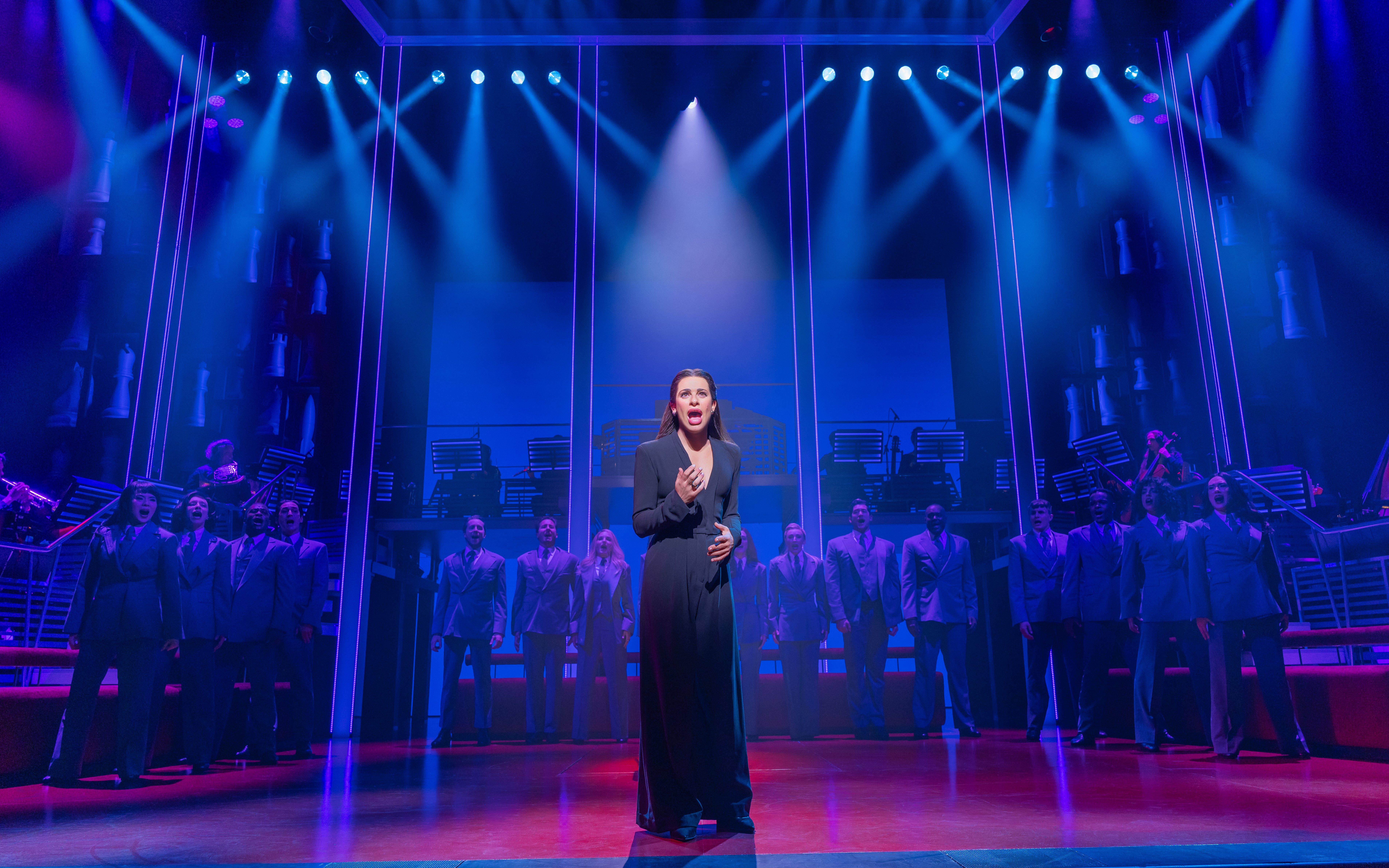 Stage performance of Chess Broadway show with lead singer and ensemble under blue lights.