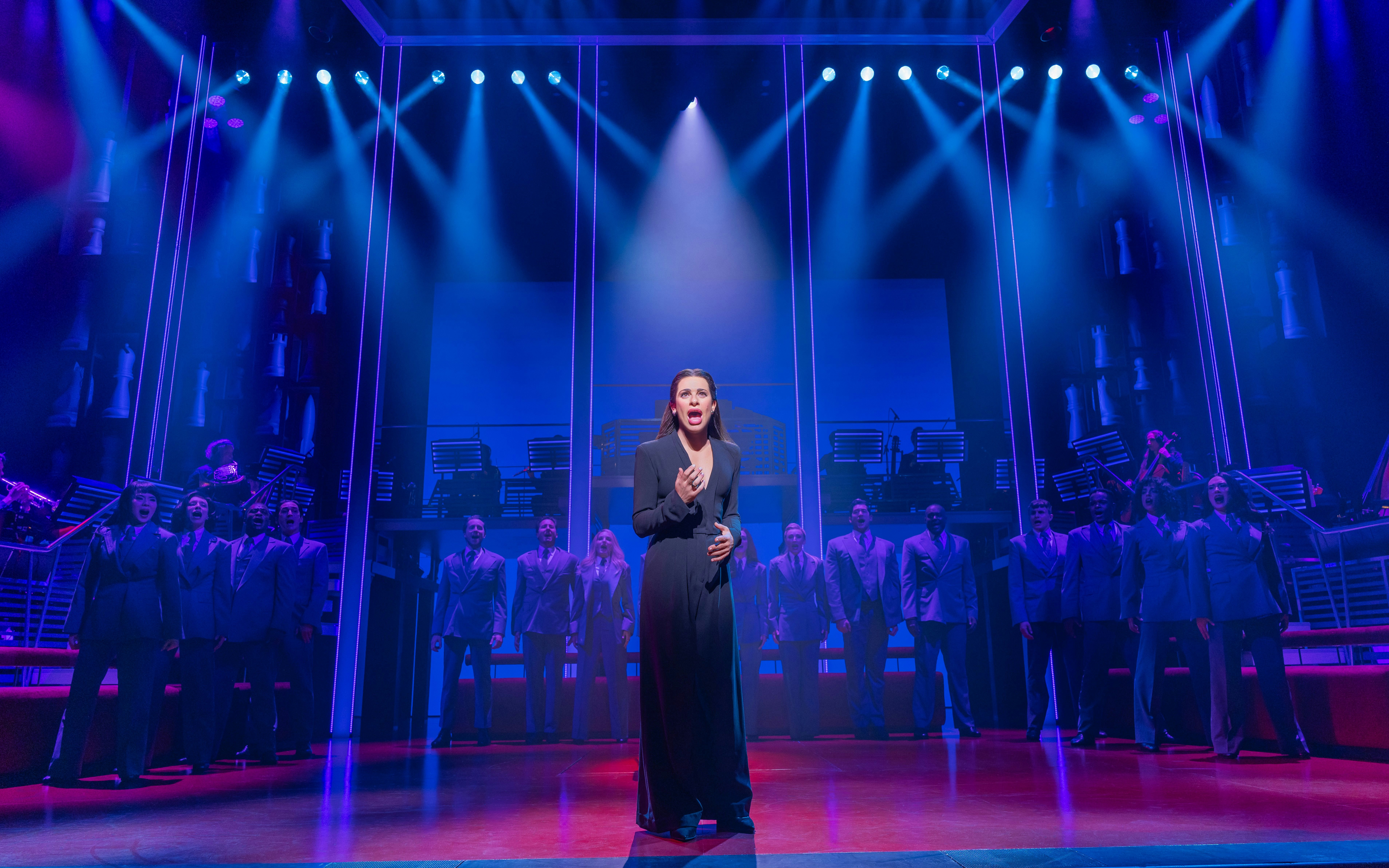 Stage performance of Chess Broadway show with lead singer and ensemble under blue lights.