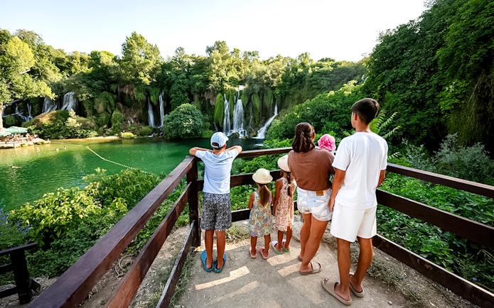 Guests viewing Kravica Waterfalls from a wooden platform surrounded by lush greenery.