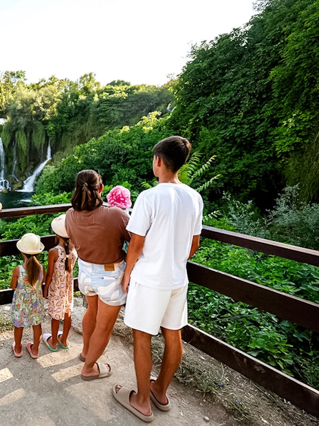 Guests viewing Kravica Waterfalls from a wooden platform surrounded by lush greenery.