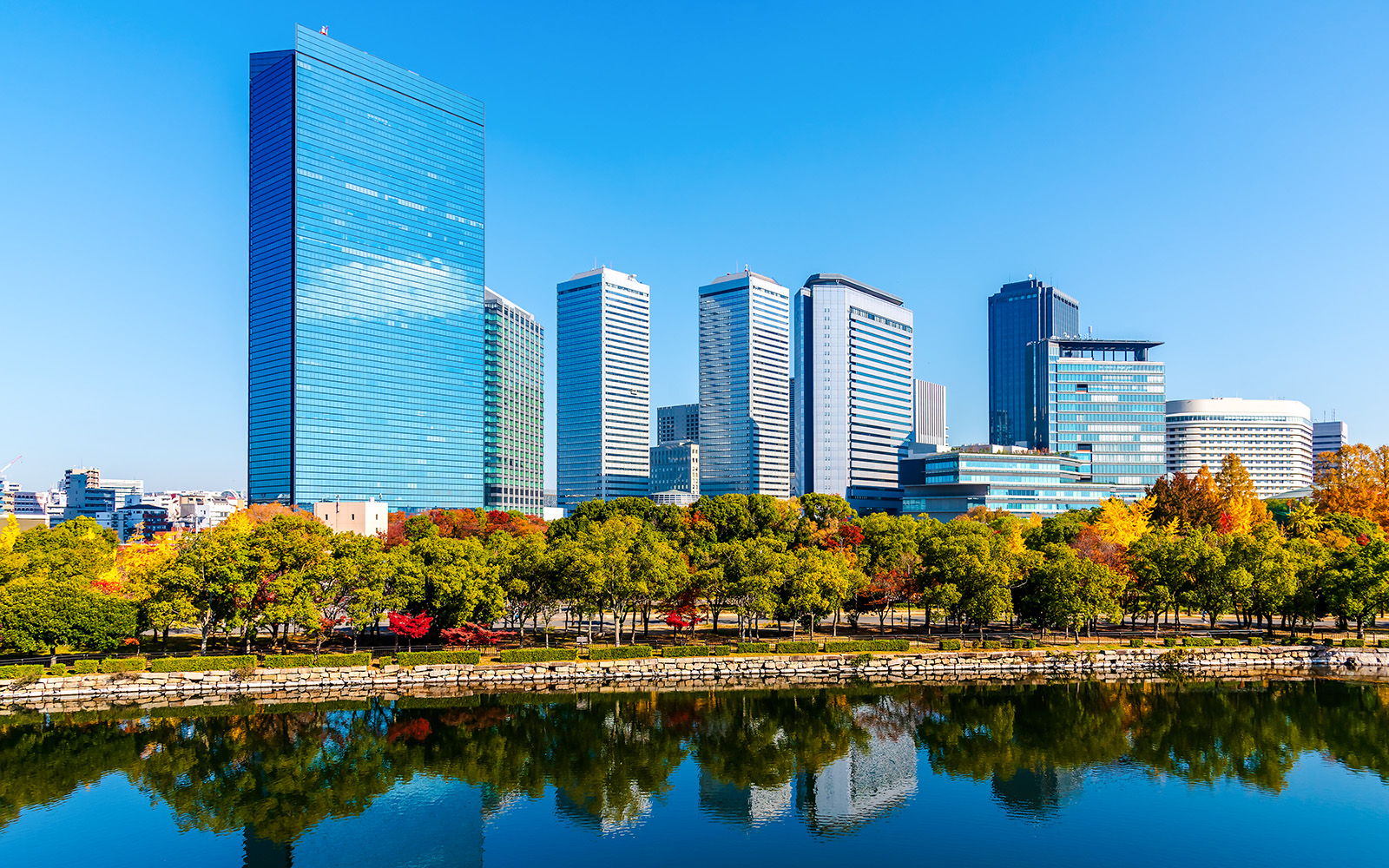Skyscrapers of Osaka Business Park reflecting in a river, surrounded by autumn trees.
