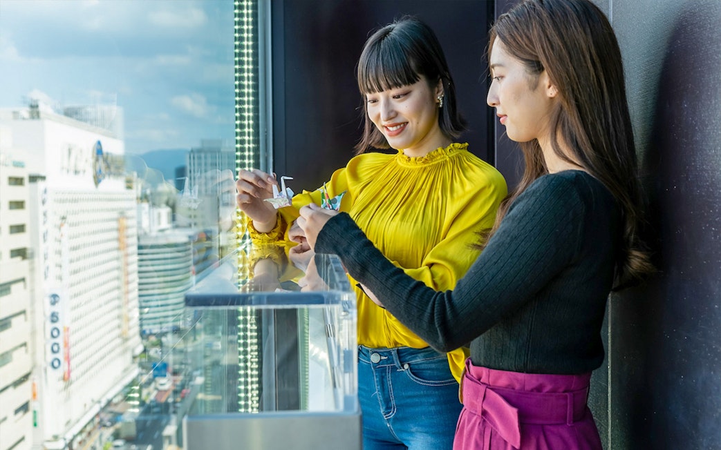 Visitors folding origami cranes at Hiroshima Orizuru Tower with city view.