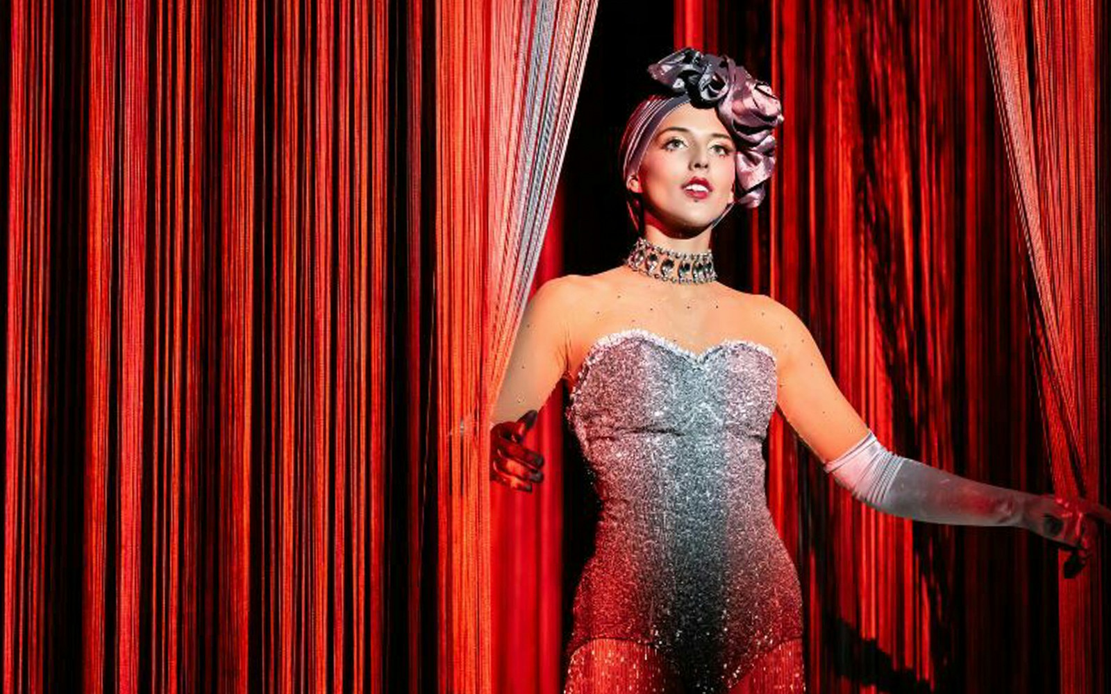 Performer in sparkling costume on stage with red curtains.