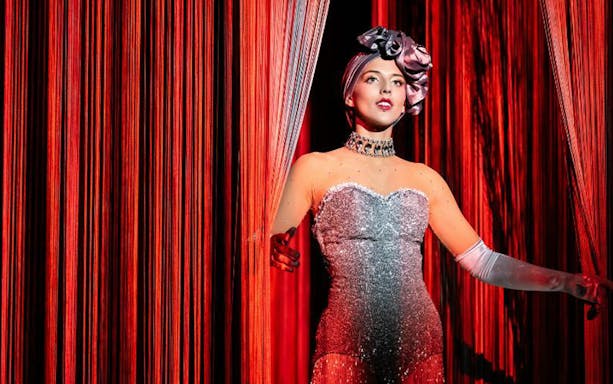Performer in sparkling costume on stage with red curtains.
