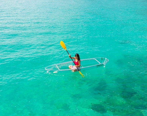 Woman kayaking in a glass-bottom kayak on clear turquoise water.