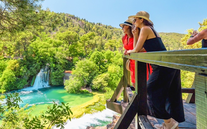 Tourists on a wooden platform observing a lush waterfall in a forest setting.