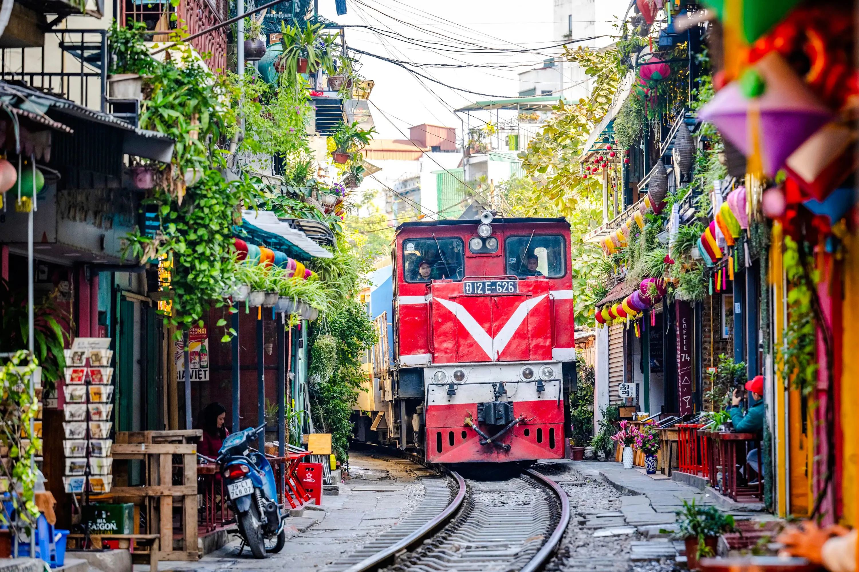 Train passing through narrow street in Hanoi, surrounded by colorful buildings and greenery.