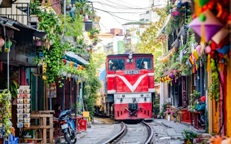 Train passing through narrow street in Hanoi, surrounded by colorful buildings and greenery.