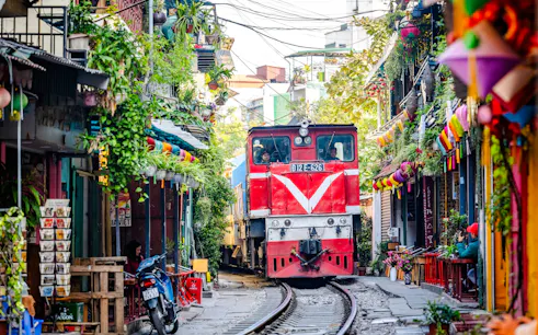 Train passing through narrow street in Hanoi, surrounded by colorful buildings and greenery.