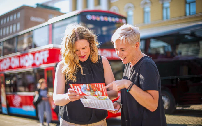 Guests checking hop on hop off bus schedule in front of a red double-decker bus.