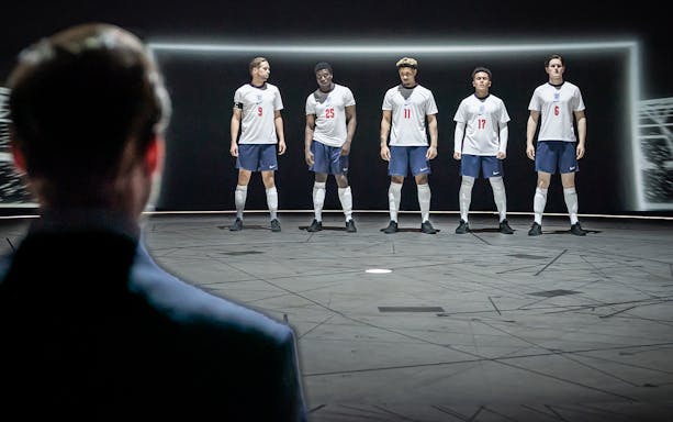Five soccer players in England jerseys stand on stage facing an audience, related to "Dear England.