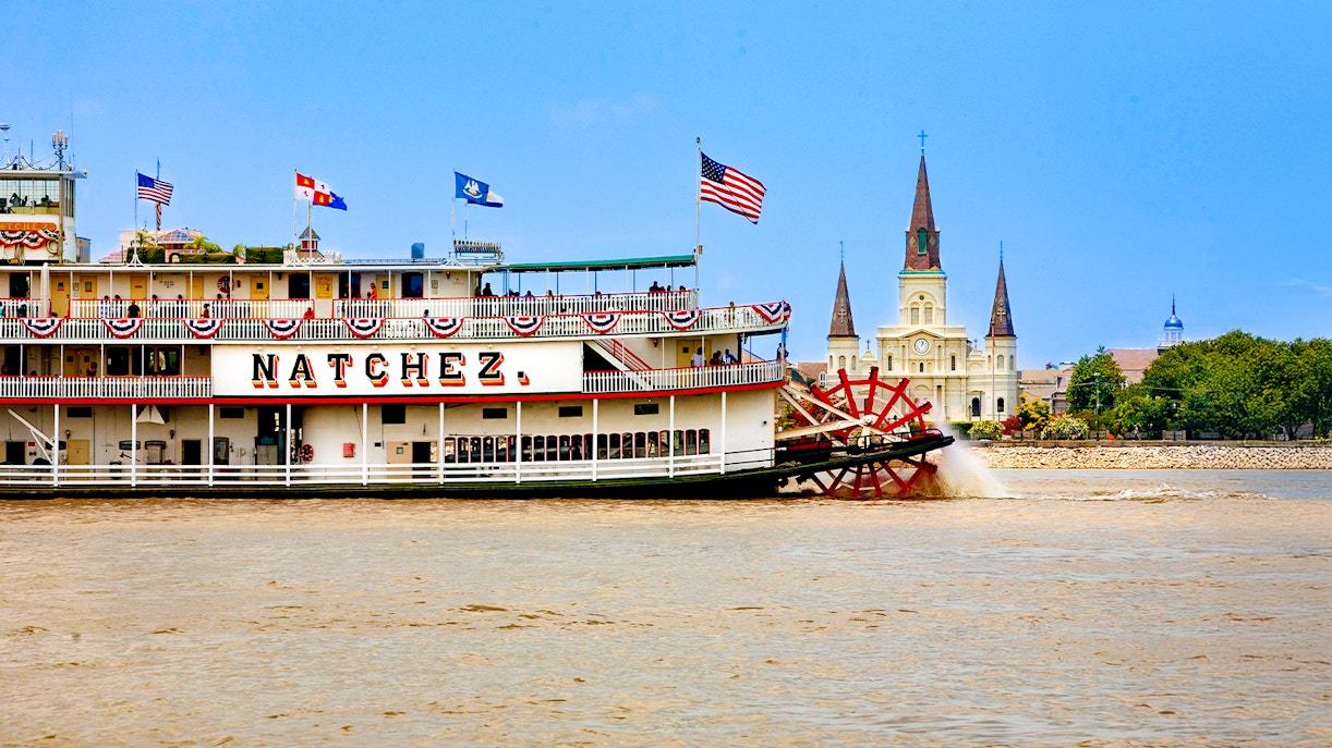 Steamboat Natchez cruising on New Orleans waters with city skyline in the background.