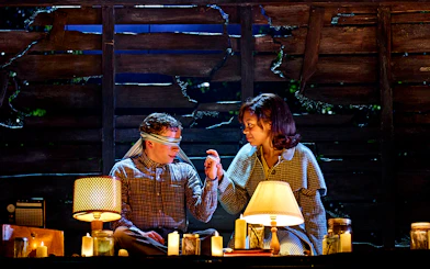 Two people in a dimly lit room with candles, one blindfolded, from "Stranger Things: The First Shadow.
