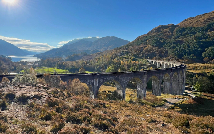 Glenfinnan Viaduct spanning lush Scottish Highlands with distant lake view.