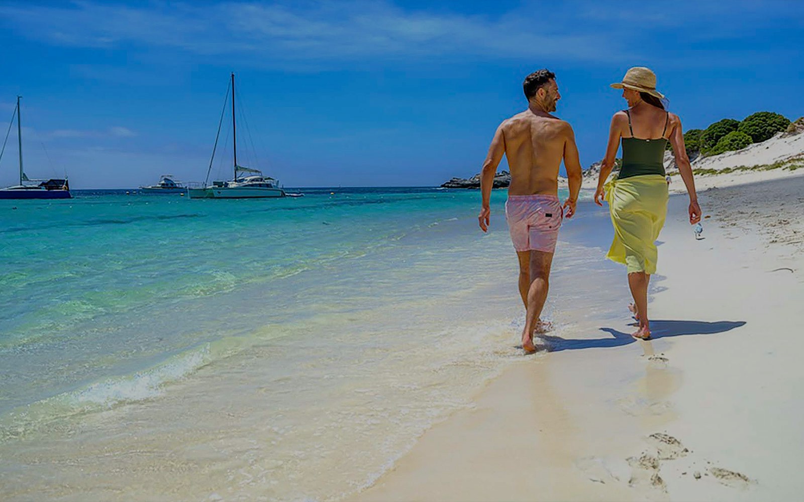 Couple walking along the beach with sailboats in the water at Rottnest Island.