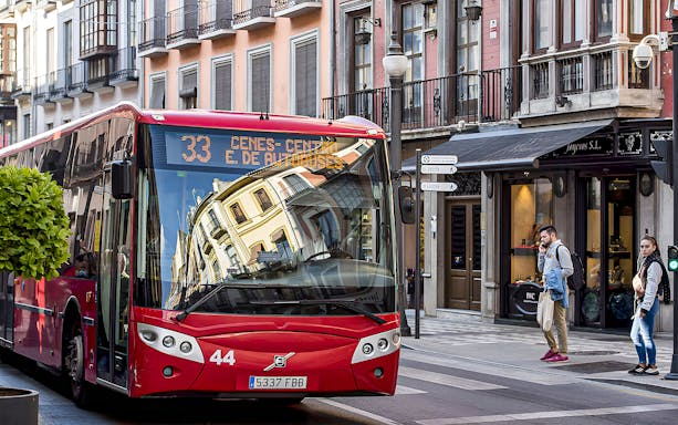 Red bus on a street in Granada, Spain, near shops and pedestrians.