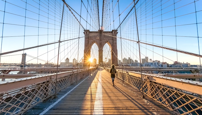 Woman walking across Brooklyn Bridge in New York City with skyline in background.